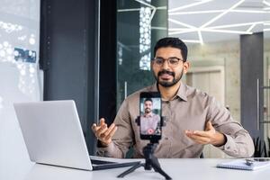 erfolgreich Geschäft Trainer Lehrer Aufzeichnung online Ausbildung Kurs, Mann Innerhalb Büro beim Arbeitsplatz mit Telefon und Stativ, Sitzung beim Schreibtisch mit Laptop. foto