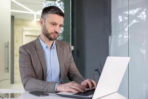 konzentriert reifen männlich Fachmann mit Computer im ein zeitgenössisch Arbeitsplatz Einstellung, Anzeigen ernst Konzentration. foto