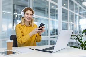 lächelnd Frau tragen Kopfhörer Arbeiten entfernt im ein modern Büro Raum, mit ein Smartphone und Laptop, genießen Fernbedienung Arbeit Leben. foto