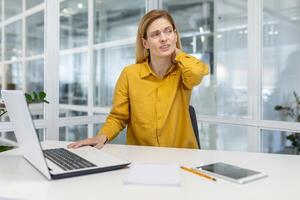 Frau im Gelb Hemd erleben Hals Schmerzen während Arbeiten beim ihr Schreibtisch im ein modern Büro Umgebung mit Laptop und Tablette auf das Tisch. foto