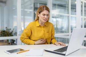 ein Frau im ein Gelb Hemd ist erleben Bauch Schmerzen während Arbeiten auf ein Laptop im ein modern Büro Umfeld. foto