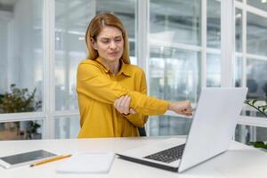 ein Frau im ein Gelb Hemd erleben Ellbogen Schmerzen während Arbeiten auf ihr Laptop im ein modern Büro Umfeld. foto