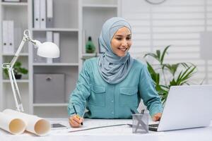 lächelnd Muslim weiblich Architekt Arbeiten mit Blaupausen und ein Laptop im ein gut beleuchtet Büro Raum, vermitteln Fokus und Professionalität. foto