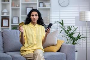 verärgert spanisch lockig behaart Mädchen Sitzung im Licht Leben Zimmer auf Sofa halten Karte und Telefon, Hindu Frau frustriert weil von gescheitert Zahlung, Betrug im online speichern, suchen beim das Kamera. foto