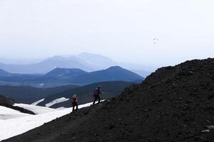 Touristen mit Rucksäcke und Trekking Stangen sind Stehen auf ein Berg suchen beim ein Gleitschirm im das Distanz. Spitzen im das Hintergrund verschwinden in das Dunst. das Schnee Lügen zwischen das Pisten foto