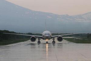 frontal Aussicht von Flugzeug Kommen im zum Landung gegen Hintergrund von schneebedeckt Berg Spitzen auf regnerisch wolkig Tag. das Licht Pfad von Scheinwerfer kriecht entlang das nass Runway gegenüber das Flughafen foto