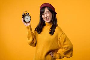 schließen oben von ein beschwingt Gelb Alarm Uhr und ein glücklich jung asiatisch Frau im ein modisch Gelb Sweatshirt und rot Baskenmütze, ausströmend ein Sinn von Pünktlichkeit und Eleganz gegen ein Gelb Hintergrund. foto