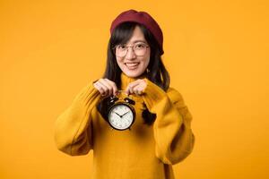 schließen oben von ein beschwingt Gelb Alarm Uhr gehaltenen durch ein jung asiatisch Frau, ausströmend Glück im ihr stilvoll Gelb Sweatshirt und rot Baskenmütze. foto