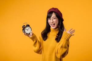 schließen oben von ein beschwingt Gelb Alarm Uhr gehaltenen durch ein glücklich jung asiatisch Frau im ein stilvoll Gelb Sweatshirt und rot Baskenmütze, betonen das Bedeutung von Zeit Management. foto
