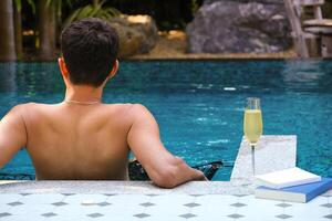 ein Mann entspannend im ein Schwimmen Schwimmbad mit ein Glas von Champagner und Buch beim ein Luxus Erholungsort. Urlaub, Reisender, entspannen Zeit Konzept. tropisch exotisch Ferien Urlaub, Tourismus und Reise. foto