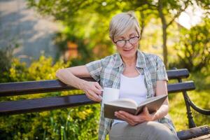 glücklich Senior Frau genießt lesen Buch und Trinken Kaffee auf ein Bank im ihr Garten. foto