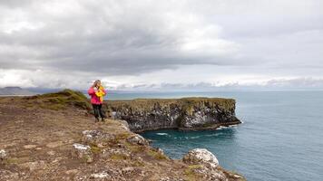 schön Mitte alt Frau Gehen entlang ein Cliff im Island tragen ein Rosa Anorak im Vorderseite von ein Blau Meer foto