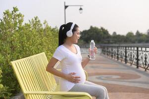 schön schwanger Frau Trinken Flasche von rein Wasser im das Park, gesund und aktiv Schwangerschaft Lebensstil Konzept. foto