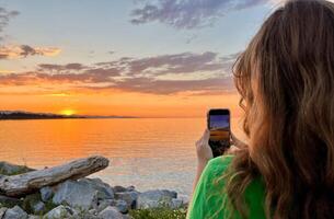 ein Teenager Mädchen im ein Grün T-Shirt schießt ein schön Sonnenuntergang auf das Pazifik Ozean sie hält ein iPhone 13 Telefon im ihr Hände blond rot Haar sichtbar Steine Rahmen Sonne Meer und Himmel foto