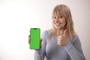 liefern Nachfrage ein Erwachsene Frau 45-50 mit blond Haar im ein Blau Kleid ist Stehen mit Handy, Mobiltelefon Telefon Grün Bildschirm Chroma Schlüssel lächelt Werbung irgendein Produkte aufrichtig Lächeln schön Gesicht verschwommen Hintergrund foto
