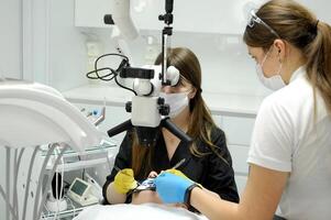 Kofferdame Klemme zum Kofferdame das Prozess von Dental Prothetik mit ein Mikroskop, weiblich Arzt im Gelb Handschuhe, geduldig Zahnarzt mit geduldig, mit Dental Aushärten Licht foto