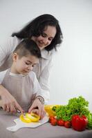 Kochen Kurse zum Kinder Mama unterrichtet zu Schnitt Gemüse mit Messer Gelb Glocke Pfeffer fein gehackt in Streifen im Familie beim Zuhause auf Weiß Hintergrund Schürze Küche Gemüse Grüner Salat Blätter foto