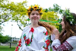 Mädchen sind Sitzung im Park warten zum Urlaub zu weben Haar flechten Zopf Kränze von Blumen auf Kopf Hilfe Freund Zärtlichkeit Schönheit jung Friseur jung Frauen sind vorbereiten zum Feier foto