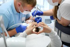 jung Mädchen zeigen Herz mit Hände Dental Büro Bürsten Zähne Bleaching Dental Verfahren Arzt und Krankenschwester Speichel Absaugen und andere neueste Technologien im Blau Handschuhe und Masken selbst Pflege Liebling foto