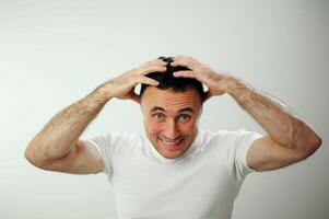 Sieg Spaß Ferien Reiseanders heiter positiv Emotionen von ein Erwachsene Mann mit grau schwarz Haar öffnen komisch heiter Blau Augen Weiß T-Shirt grau Hintergrund Falten Stirn Stock aus Zunge foto