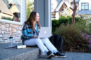 Teenager Mädchen Sitzung auf Veranda im Hände mit Telefon sie hat Weiß Jeans Blau Bluse Nächster zu Rucksack mit Wasser Privat Sektor Blumen Frühling Sommer- Schule Ausbildung warten zum Eltern vergessen Schlüssel foto