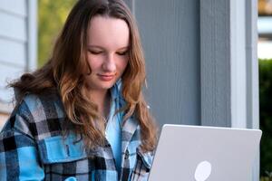 Teenager Mädchen mit ein Laptop Sitzung auf das Veranda von das Haus. sie Aufpassen das Nachrichten tun Hausaufgaben online Konferenz Rucksack Mappe Wasser Flasche Bleistift gewöhnliche Menschen echt Leben foto