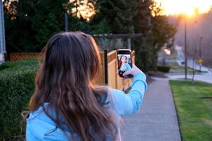 ein jung Frau nimmt ein Selfie auf Straße während Sonnenuntergang ein Teenager Mädchen im ihr Hände mit ein Telefon in der Nähe von das Haus und ein Zaun Privat Sektor Hintergrundbeleuchtung können Sein gesehen reflektiert im das Gadget Monitor Bildschirm foto