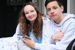 Teenager Junge und Mädchen lächelnd und halten Handys Jungs Mädchen auf das Veranda mit ein Aktentasche eingewickelt im ein Decke suchen beim das Telefon Fotos horizontal Position Jugendliche