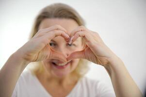 ein Frau zeigt an ihr Herz mit ihr Hände ein heiter aussehen durch ihr Hände beim das Kamera Weiß Hintergrund Interesse Spaß Freude angenehm Emotionen Glück foto