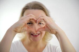 mittleren Alters Frau zeigen Herz mit Hände sieht aus durch Loch lächelt Liebe Erwartungen Anerkennung Zärtlichkeit Schönheit Hände im Schärfe selektiv Schärfe auf Weiß Hintergrund weiblich Gefühle foto