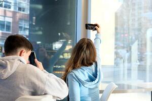 Leben von Jugendliche auf Telefon Junge und Mädchen Sitzung im Cafe mit Rücken zu Kamera Mädchen macht Selfie Junge geschlossen selbst mit Telefon Sozial Netzwerke Profil Leben im Sozial Netzwerke verbringen Zeit zusammen Datum foto