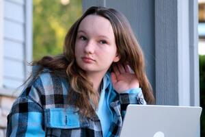 Teenager Mädchen Sitzung auf Veranda im Hände mit Telefon sie hat Weiß Jeans Blau Bluse Nächster zu Rucksack mit Wasser Privat Sektor Blumen Frühling Sommer- Schule Ausbildung warten zum Eltern vergessen Schlüssel foto