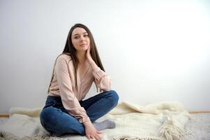 ein Mädchen im Jeans und ein Rosa Bluse sitzt auf das Fußboden auf ein Plaid sieht aus in das Rahmen lange Haar auf ein Weiß Hintergrund Ruhe aussehen foto