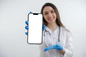 Telefon mit ein Weiß Bildschirm Mädchen Arzt zeigt an ein Hand im Blau Handschuhe auf ein leeren Raum zum Text Weiß Hintergrund Ihre Werbung ist schön süß Frau lächelnd verschwommen auf Hintergrund Nahansicht Gadget foto