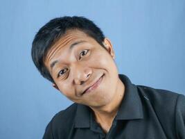 komisch Gesicht Ausdruck von jung asiatisch Mann lächelnd suchen beim Kamera, isoliert auf Blau Hintergrund. foto