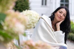 ein Frau eingewickelt im ein Sahne Schal sitzt inmitten blüht, ihr Inhalt Lächeln reflektieren Selbstakzeptanz und Anmut beim Lebensmitte inmitten hormonell Übergang. foto