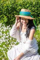 lächelnd Frau im Weiss, Stroh Hut mit Blau Band, inmitten ein Grün und Weiß Gänseblümchen Feld, froh Sommer- Stimmung, Natur umarmen, sonnig Hintergrund. foto