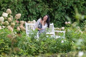 ruhen auf ein Park Bank, ein Frau mit ein Inhalt lächeln, umgeben durch dicht Hortensien, verkörpert entspannt Eleganz im Naturen umarmen. foto