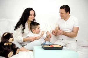 verbringen Zeit mit Familie im Weiß Kleider spielen mit Essstäbchen von orientalisch Essen hölzern Stöcke Trommel Lachen haben Spaß im Bett zusammen Essen Sushi Sanft Spielzeug braun Teddy Bär Tag aus Papa Mama Sohn foto