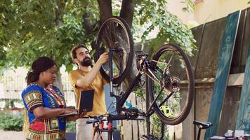 gesund schwarz Frau suchen auf Minicomputer zu helfen jung Mann Reparatur beschädigt Fahrrad. Freund und Freundin Aufrechterhaltung Fahrrad Rad während nutzen Laptop zum jährlich Sommer- draussen Radfahren. foto