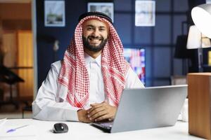 männlich Person gekleidet im traditionell Arabisch Kleidung ist sitzend mit seine Laptop. zuversichtlich und technisch versiert, das arabisch Mann lächelt beim das Kamera, verkörpern das unternehmerisch Geist von das Digital Alter. foto