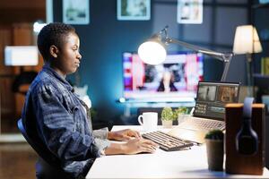 schwarz Frau sitzt beim ihr Schreibtisch, Verwendet ein Laptop, und Typen auf das Klaviatur. sie ist vollständig absorbiert im ihr arbeiten, Surfen und Bearbeitung Inhalt und demonstrieren ihr Kreativität und Produktivität. foto