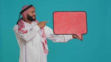 Muslim Mann zeigt an Rede Blase auf Kamera, Erstellen Werbung zum Neu Netz Werbung. jung Kerl Arbeiten im Marketing Industrie mit isoliert Karton Zeichen, leeren Werbetafel. foto