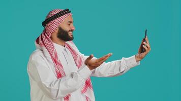 islamisch Kerl Gruß freunde auf online Forderung, winken und fangen oben mit Menschen auf Fernbedienung kommunizieren Netzwerk. Mitte östlichen Mann im National Muslim Kleider reden auf Videokonferenz. foto