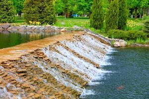 das Wasser eines schönen dekorativen Wasserfalls fließt schnell in einen Sommerpark.