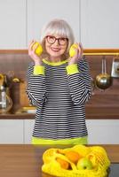 Fröhliche hübsche ältere lächelnde Frau in gestreiftem Pullover, die Zitronen für Limonade hält, während sie in der Küche steht. gesunder, saftiger Lebensstil, Zuhause, Seniorenkonzept. foto