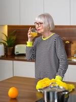 fröhliche hübsche ältere lächelnde Frau in gestreiftem Pullover, die Orangensaft trinkt, während sie in der Küche steht. gesunder, saftiger Lebensstil, Zuhause, Seniorenkonzept.