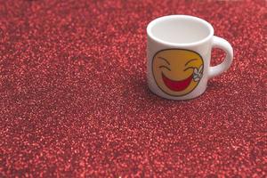 weiße Tasse mit einem lächelnden Emoticon auf einem rot glitzernden Hintergrund. Tag des Lächelns. Platz kopieren. foto