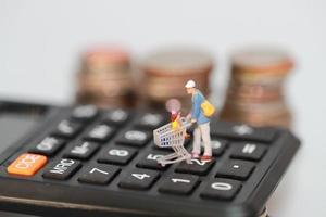 Shopper und Trolley laufen auf einem Taschenrechner mit unscharfen Münzen dahinter als Hintergrund foto