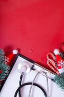 flache Zwischenablage mit medizinischem Stethoskop, Brille, Weihnachtsbaum auf rotem Hintergrund mit Kopierraum foto